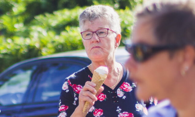 Starija žena u cvjetnoj majici uživa u sladoledu na ljetnom suncu, dok hoda ulicom uz parkirane automobile – spontani trenutak iz svakodnevice.