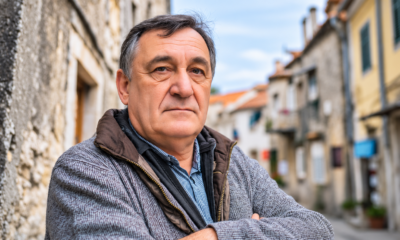 Stariji muškarac ozbiljnog izraza lica stoji prekriženih ruku u uskoj kamenoj ulici, djelujući nezadovoljno ili zamišljeno, u toploj zimskoj jakni i sivom puloveru.