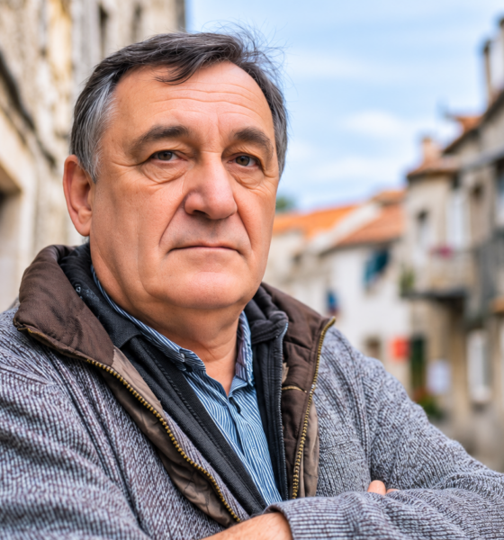 Stariji muškarac ozbiljnog izraza lica stoji prekriženih ruku u uskoj kamenoj ulici, djelujući nezadovoljno ili zamišljeno, u toploj zimskoj jakni i sivom puloveru.