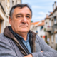 Stariji muškarac ozbiljnog izraza lica stoji prekriženih ruku u uskoj kamenoj ulici, djelujući nezadovoljno ili zamišljeno, u toploj zimskoj jakni i sivom puloveru.