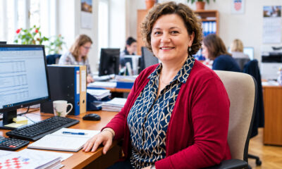 Nasmiješena pedesetogodišnja računovotkinja u crvenoj vesti sjedi za radnim stolom ispred računala u uredu.