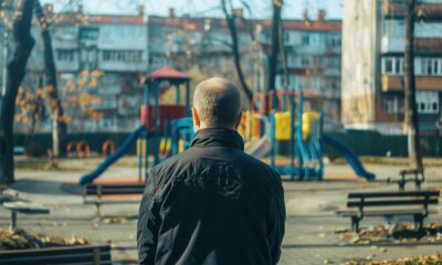Stariji muškarac u crnoj jakni gleda prema praznom dječjem igralištu ispred stambenih zgrada.