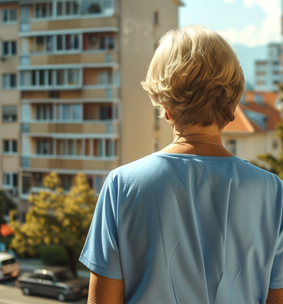 Starija žena u plavoj majici gleda u daljinu prema zgradi doma za starije osobe u kojem je smještena njezina majka. Njezina leđa i pognuta glava odražavaju brigu i tugu dok razmišlja o majčinom iskustvu unutar doma.