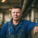 Muškarac u plavom radnom odijelu stoji u industrijskoj hali, naslonjen na metalnu ogradu, s ozbiljnim izrazom lica. U pozadini se naziru strojevi i dugačak hodnik tvornice.