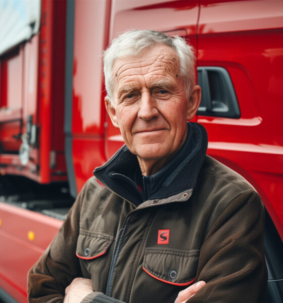 Stariji vozač kamiona stoji ispred crvene Scania S700 šleper prikolice, nasmijan i s prekriženim rukama.