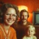 **SEO meta opis fotografije:** Obiteljski portret iz 1970-ih prikazuje nasmijanu ženu s velikim naočalama, muškarca s brkovima i dijete ispred starog televizora u retro dnevnoj sobi, obasjanoj toplim svjetlom lampe.
