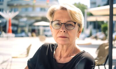Starija žena kratke plave kose i naočala sjedi na otvorenoj terasi kafića, odjevena u crnu bluzu, ozbiljnog izraza lica, s modernim gradskim trgom u pozadini.