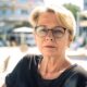 Starija žena kratke plave kose i naočala sjedi na otvorenoj terasi kafića, odjevena u crnu bluzu, ozbiljnog izraza lica, s modernim gradskim trgom u pozadini.