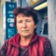 Starija žena kratke tamnocrvene kose stoji naslonjena na zid pored bankomata, odjevena u bordo prsluk i šarenu bluzu, s ozbiljnim izrazom lica na gradskoj ulici.