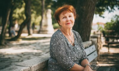 Starija žena crvene kose sjedi nasmiješena na klupi u sjenovitom gradskom parku, odjevena u crno-bijelu haljinu s uzorkom i noseći jednostavan srebrni nakit.