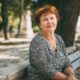 Starija žena crvene kose sjedi nasmiješena na klupi u sjenovitom gradskom parku, odjevena u crno-bijelu haljinu s uzorkom i noseći jednostavan srebrni nakit.