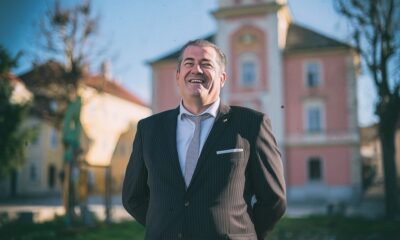Muškarac u tamnom odijelu i kravati nasmijano pozira ispred gradske zgrade na sunčan dan – službeni portret političara na javnoj dužnosti u urbanom okruženju.