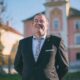 Muškarac u tamnom odijelu i kravati nasmijano pozira ispred gradske zgrade na sunčan dan – službeni portret političara na javnoj dužnosti u urbanom okruženju.