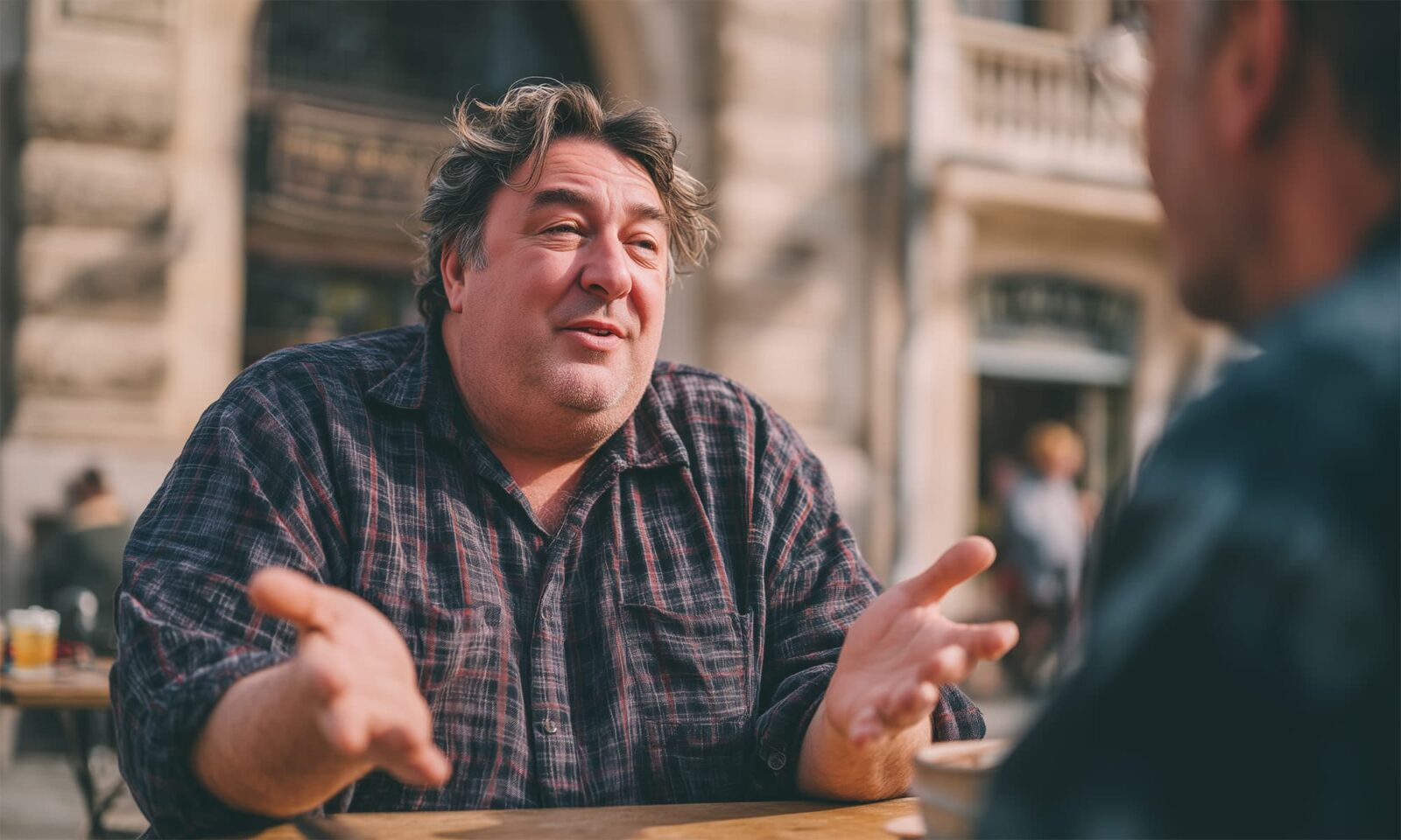 Muškarac sjedi na terasi kafića, energično gestikulira i razgovara s osobom nasuprot sebe. Izraz lica mu je živahan i pun entuzijazma, kao da objašnjava nešto važno ili zabavno. U pozadini se vidi gradska arhitektura i prolaznici u pokretu.