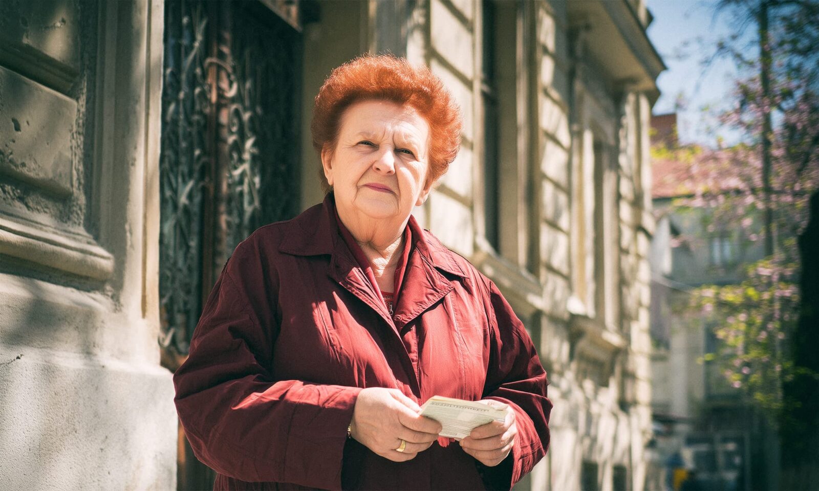 Na slici je starija žena u bordo kaputu koja stoji na sunčanom gradskom pločniku, ispred klasične kamene zgrade. U rukama drži papire ili račune, a izraz lica joj je ozbiljan, s dozom odlučnosti. Svjetlo i sjene na zidu dodatno naglašavaju atmosferu svakodnevice.