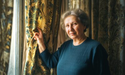 Starija žena sijede kose i tamne majice stoji kraj prozora, pridržava raskošnu zlatno-plavu zavjesu i gleda mirno prema kameri u toplom, kućnom ambijentu.