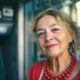 Našminkana starija žena s razbarušenom kosom, šarenim naušnicama i ogrlicom od crvenih kuglica stoji nasmijana ispred bankomata na gradskoj ulici, odjevena u crvenu ljetnu haljinu.