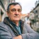 Stariji muškarac ozbiljnog izraza lica stoji prekriženih ruku u uskoj kamenoj ulici, djelujući nezadovoljno ili zamišljeno, u toploj zimskoj jakni i sivom puloveru.