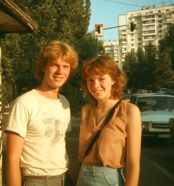 Autentična scena iz Jugoslavije 1980-ih: mladi par nasmijanih lica pozira na prometnoj ulici, okružen starim automobilima i oglasima iz novina – sjećanje na bezbrižno doba.