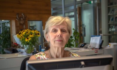 Starija žena sjedi za računalom na recepciji moderno uređenog ureda, okružena dokumentima i buketom sunovrata, zračeći profesionalnošću i ozbiljnošću.