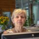 Starija žena sjedi za računalom na recepciji moderno uređenog ureda, okružena dokumentima i buketom sunovrata, zračeći profesionalnošću i ozbiljnošću.