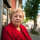 Starija žena u crvenom kaputu stoji pokraj bankomata, ozbiljnog izraza lica, simbolično prikazujući iščekivanje mirovine i svakodnevne financijske brige umirovljenika.