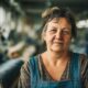 Starija žena u radnoj odjeći stoji u tekstilnoj tvornici među rolama tkanine, simbolizirajući dugogodišnji rad i iskustvo u industriji.