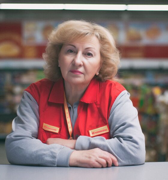 Starija žena u crvenom prsluku trgovkinje sjedi za pultom u trgovini, s ozbiljnim izrazom lica, okružena policama s robom.