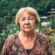 Starija žena stoji u svom dvorištu s kokošima u pozadini, odjevena u bluzu s uzorkom, ozbiljnog izraza lica, s gustim zelenilom i gospodarskim objektima iza nje.