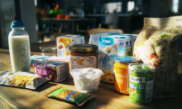 Zdravi prehrambeni proizvodi raspoređeni na drvenom stolu u kuhinji, uključujući biljna mlijeka, grickalice bez glutena, fermentirane proizvode i namaze bez aditiva, u ambalaži s oznakama prirodnih sastojaka. Sunčeva svjetlost obasjava stol, stvarajući toplu i privlačnu atmosferu.