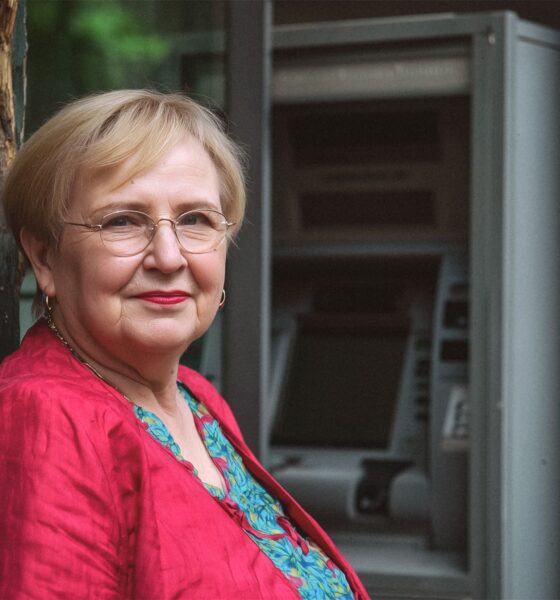 Zrela žena kratke svijetlosmeđe kose stoji naslonjena na kameni zid, gledajući ravno u kameru s blagim osmijehom. U pozadini se nazire bankomat na kojem podiže mirovinu.