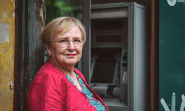 Zrela žena kratke svijetlosmeđe kose stoji naslonjena na kameni zid, gledajući ravno u kameru s blagim osmijehom. U pozadini se nazire bankomat na kojem podiže mirovinu.