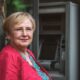 Zrela žena kratke svijetlosmeđe kose stoji naslonjena na kameni zid, gledajući ravno u kameru s blagim osmijehom. U pozadini se nazire bankomat na kojem podiže mirovinu.