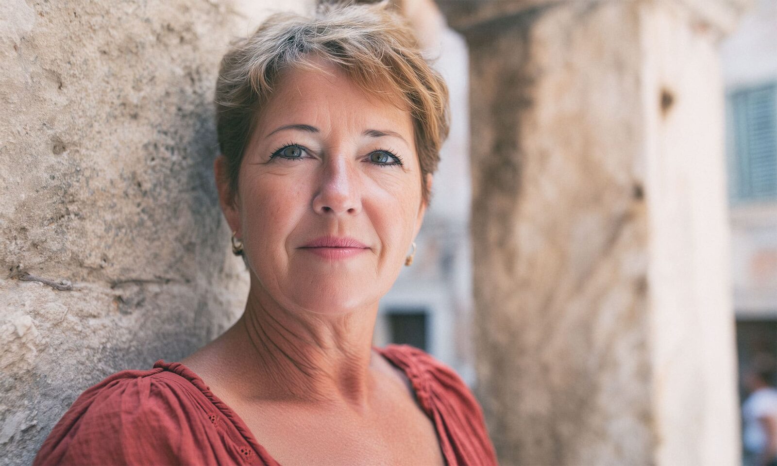 Zrela žena kratke svijetlosmeđe kose stoji naslonjena na kameni zid, gledajući ravno u kameru s blagim osmijehom. U pozadini se nazire mediteransko kameno zdanje.