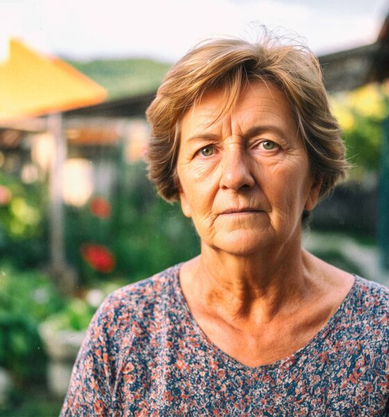 Starija žena stoji u dvorištu obiteljske kuće na selu. Iza nje se nazire vrt.