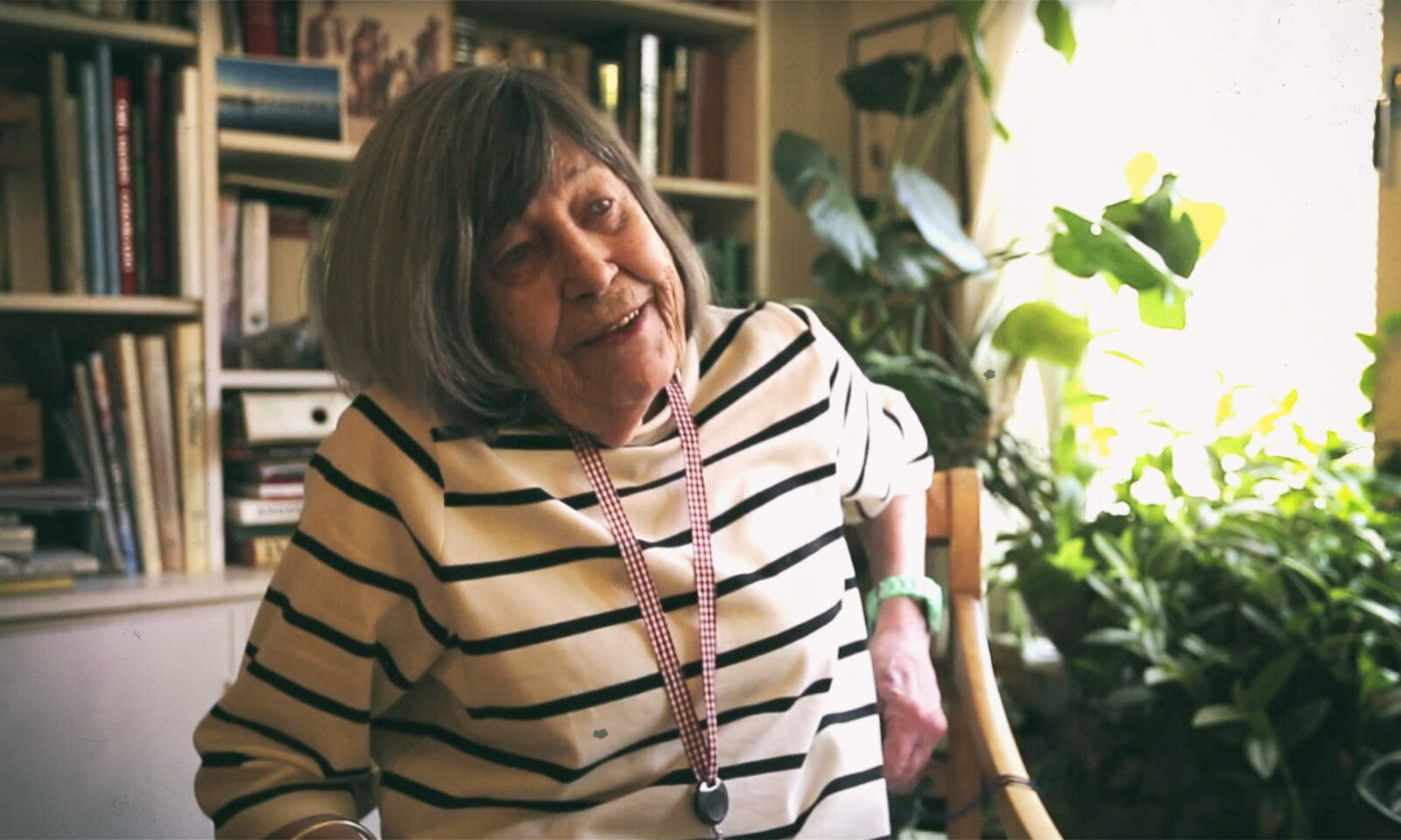 Margareta Magnusson u prugastoj majici sjedi u dnevnom boravku punom knjiga i biljaka, nasmiješena i opuštena, portret u prirodnom svjetlu