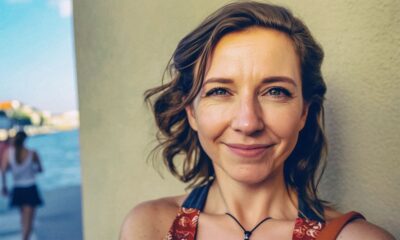 Žena srednjih godina stoji naslonjena na zid i gleda u kameru s blagim osmijehom. U pozadini se nazire more i prolaznica na šetnici, uz ljetno, sunčano ozračje.