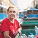Starija žena u crvenoj majici stoji pokraj kontejnera s plastičnim bocama, u pozadini plavi tramvaj u Zagrebu.