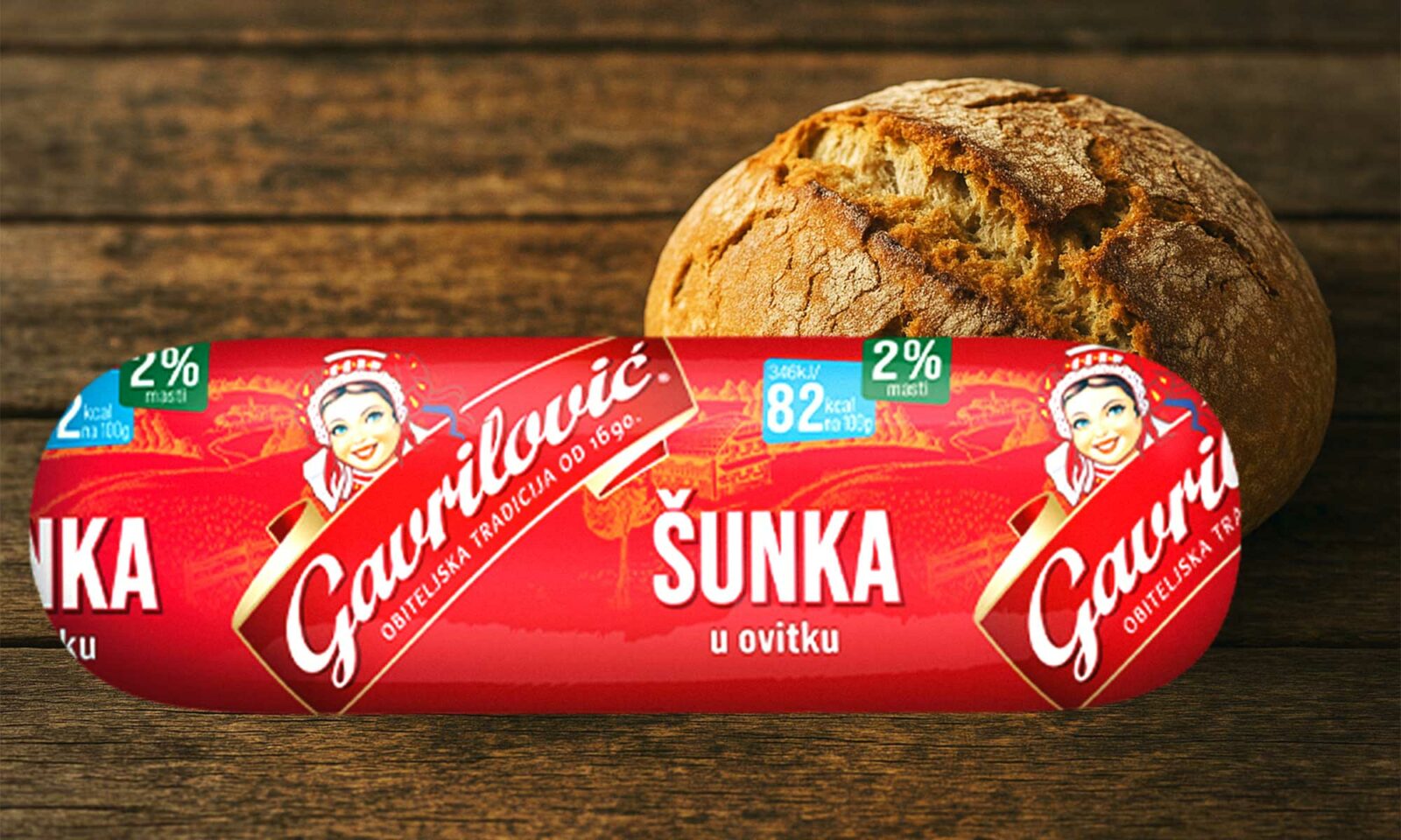 Gavrilović šunka u ovitku s oznakom 2 posto masti, ispred svježe pečenog kruha na drvenoj podlozi.