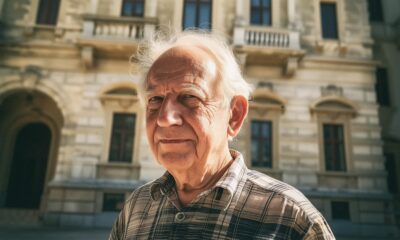 Stariji muškarac u kariranoj košulji stoji ispred kamene zgrade s velikim prozorima.