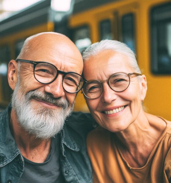 Našmiješeni stariji par s naočalama pozira ispred žutog tramvaja na stanici.