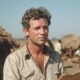 Filmski umjetnički direktor 1980-ih na setu u Africi, u pustinji ispred koliba, s bilježnicom u ruci i ozbiljnim izrazom lica.