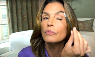 Cindy Crawford u ljubičastoj košulji češlja obrve malom četkicom u spavaćoj sobi s prozorom i zavjesama u pozadini.