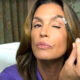 Cindy Crawford u ljubičastoj košulji češlja obrve malom četkicom u spavaćoj sobi s prozorom i zavjesama u pozadini.