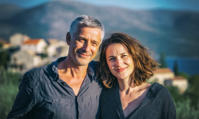 Našmiješeni par srednjih godina pozira na suncu u mediteranskom krajoliku, s kamenom arhitekturom i planinama u pozadini.