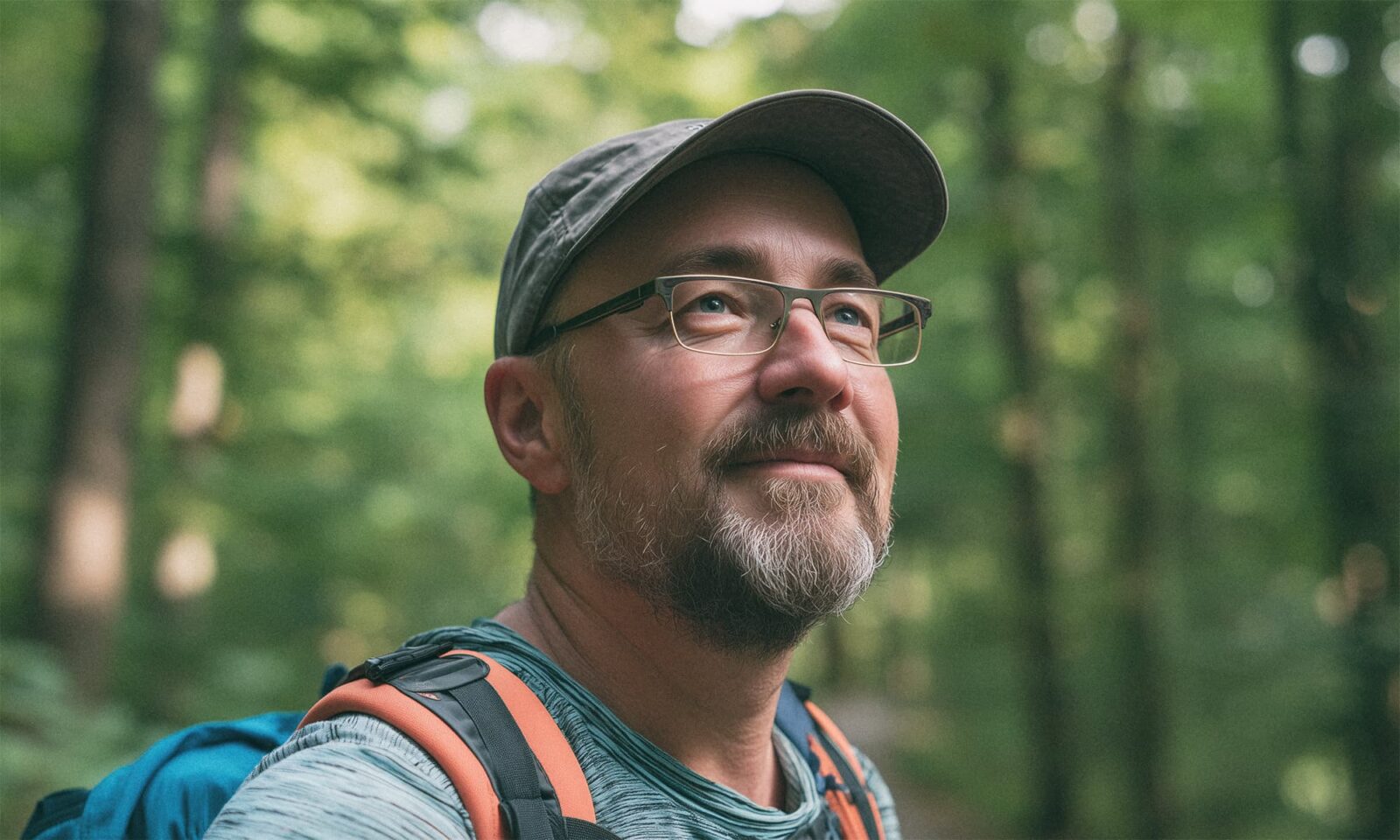 Zadovoljni sredovječni muškarac s naočalama i kapom stoji u šumi s ruksakom na leđima, s blagim osmijehom na licu i pogledom uprtim prema nebu.