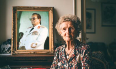 Starija žena sijede kose sjedi u dnevnom boravku uz veliki portret Josipa Broza Tita u bijeloj maršalskoj uniformi, nostalgična atmosfera.