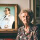 Starija žena sijede kose sjedi u dnevnom boravku uz veliki portret Josipa Broza Tita u bijeloj maršalskoj uniformi, nostalgična atmosfera.