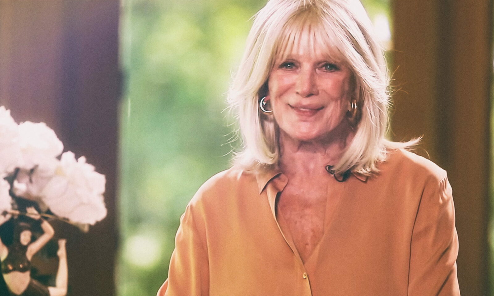Američka glumica Linda Evans, plave kose i u elegantnoj košulji boje marelice, nasmiješeno pozira u svijetlom interijeru s cvijećem u pozadini.