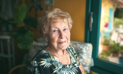 Starija žena sjedi u dnevnom boravku kraj prozora, s blagim osmijehom i u cvjetnoj haljini, prirodno osvijetljena.